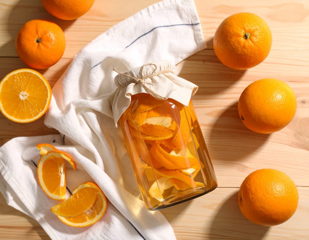 Glasgefäß mit eingelegten Zitronen- und Orangenschalen in Essig – nachhaltiger DIY-Reiniger auf einem Holztisch, umgeben von frischen Zitrusfrüchten und Sprühflasche.